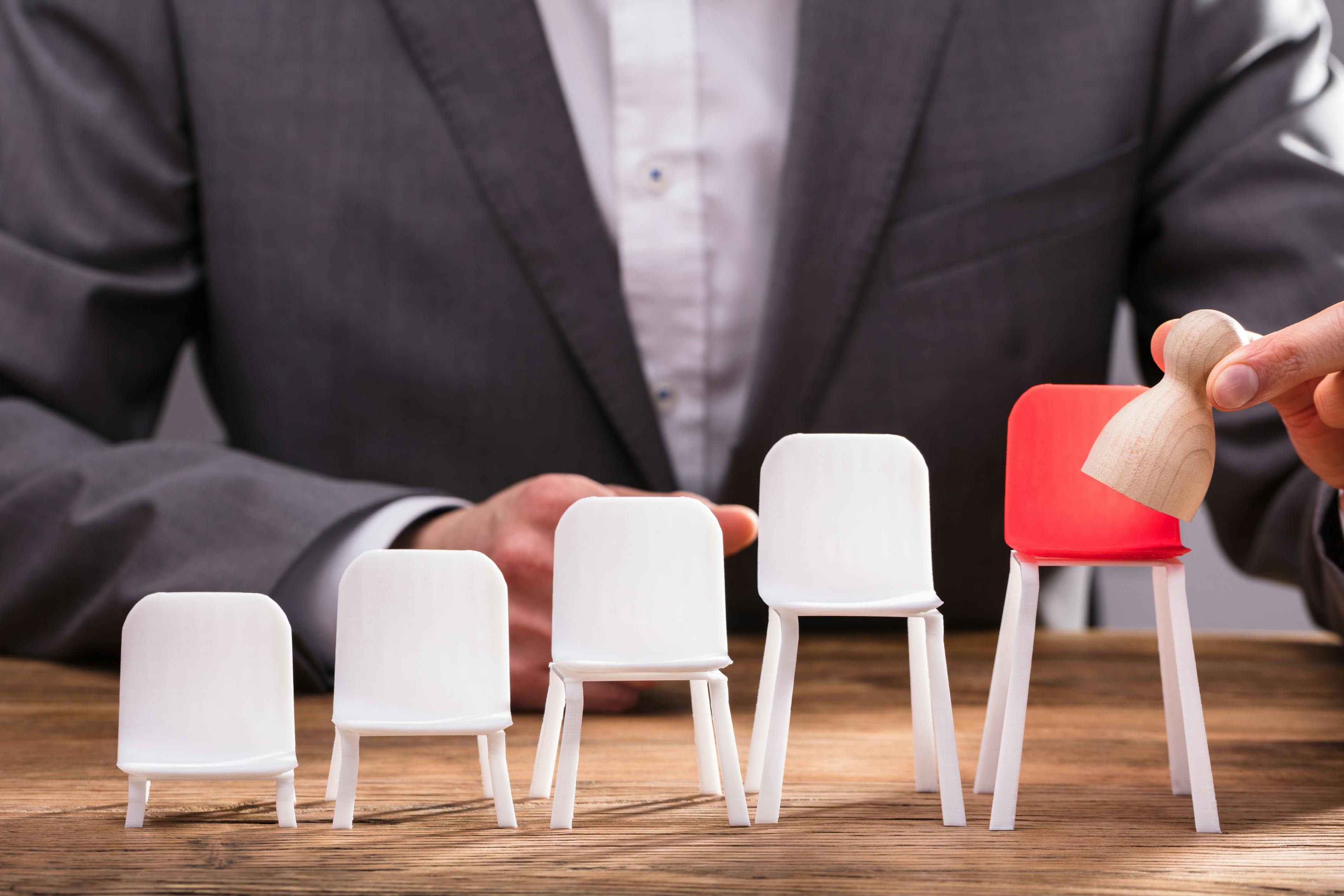 Close-up Of Businessperson`s Finger Placing Pawn On Red Chair Over Wooden Desk