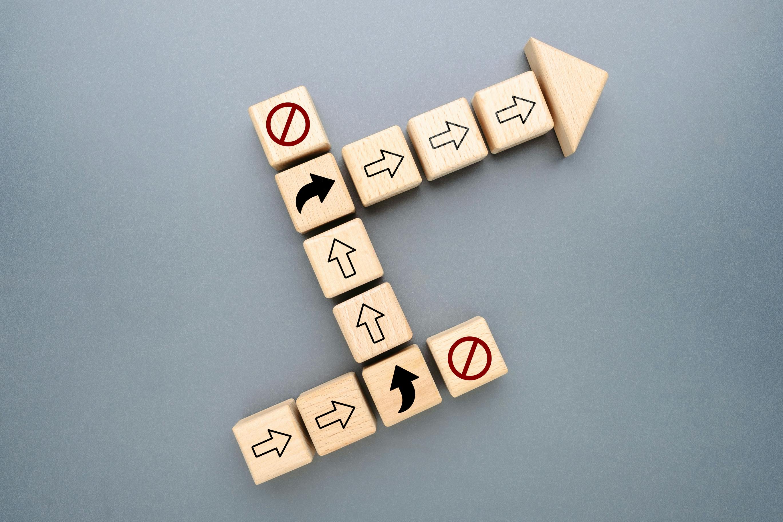 Wooden blocks with directional arrows changing path to avoid blocked routes, representing strategic pivot, business adaptation, problem solving, and navigating challenges toward success