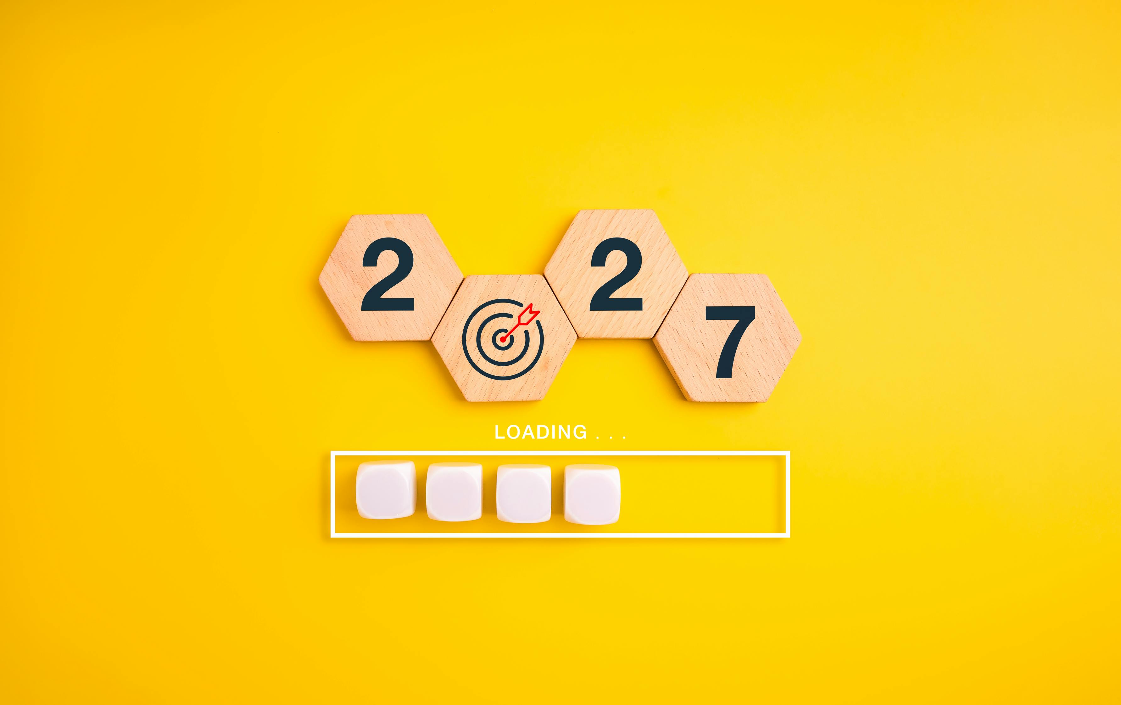 Wooden hexagon blocks form 2027 with bullseye replacing zero above loading bar on a yellow background. Preparation, progress tracking, strategic goals and readiness for the new year concept