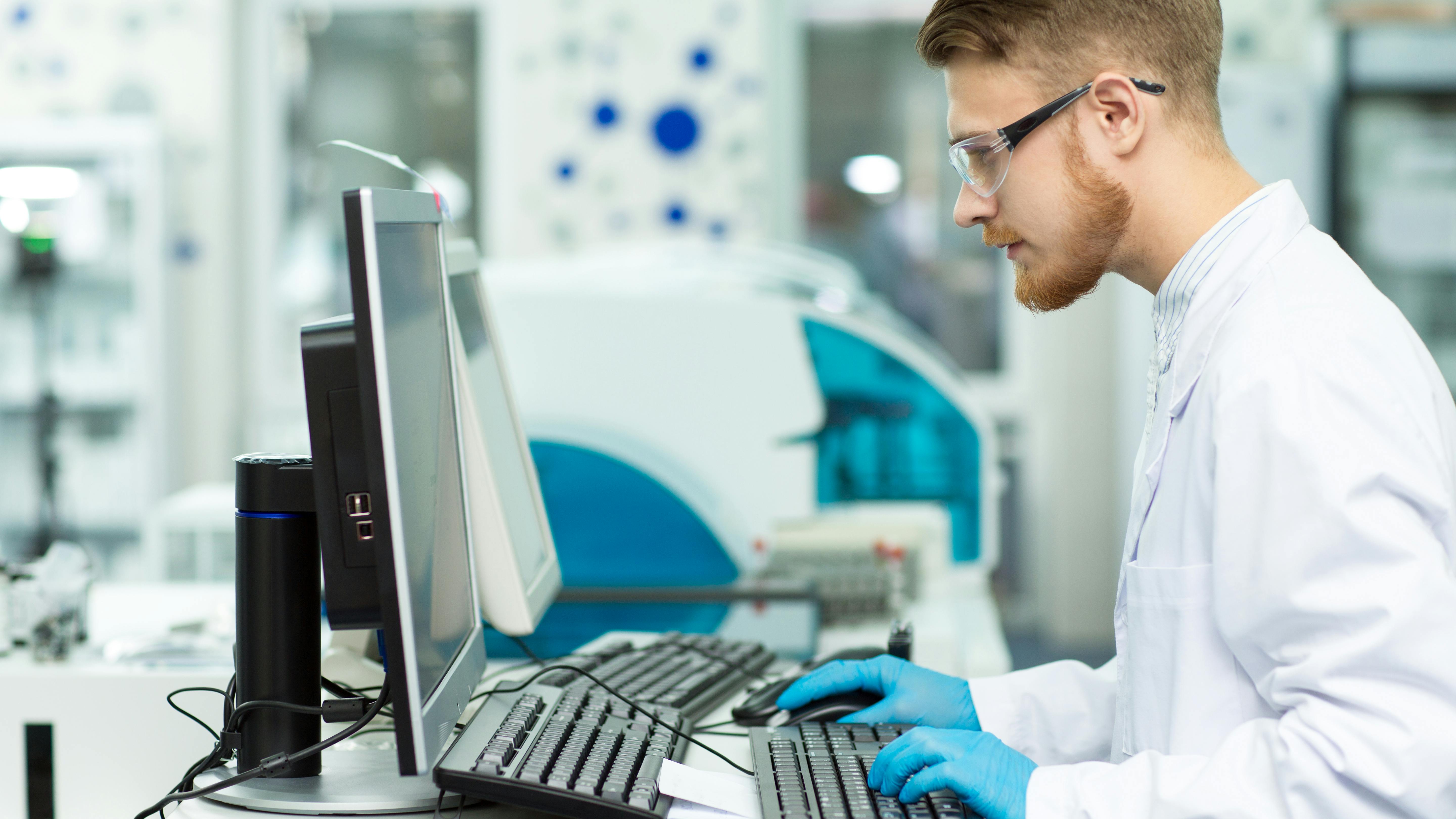 A man in a lab coat works in a computer in a healthcare lab setting.