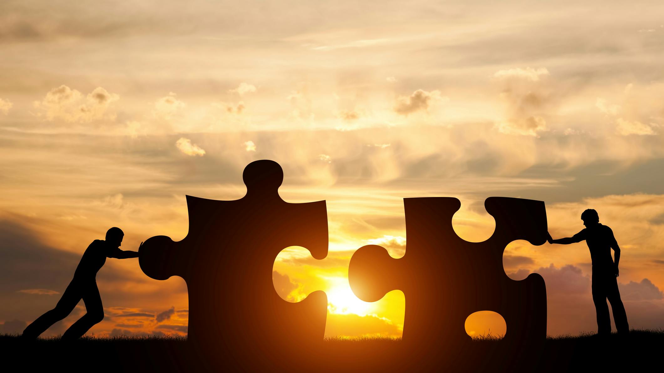 Concept of solving a problem with a silhouette of two men connecting puzzle pieces at sunset.
