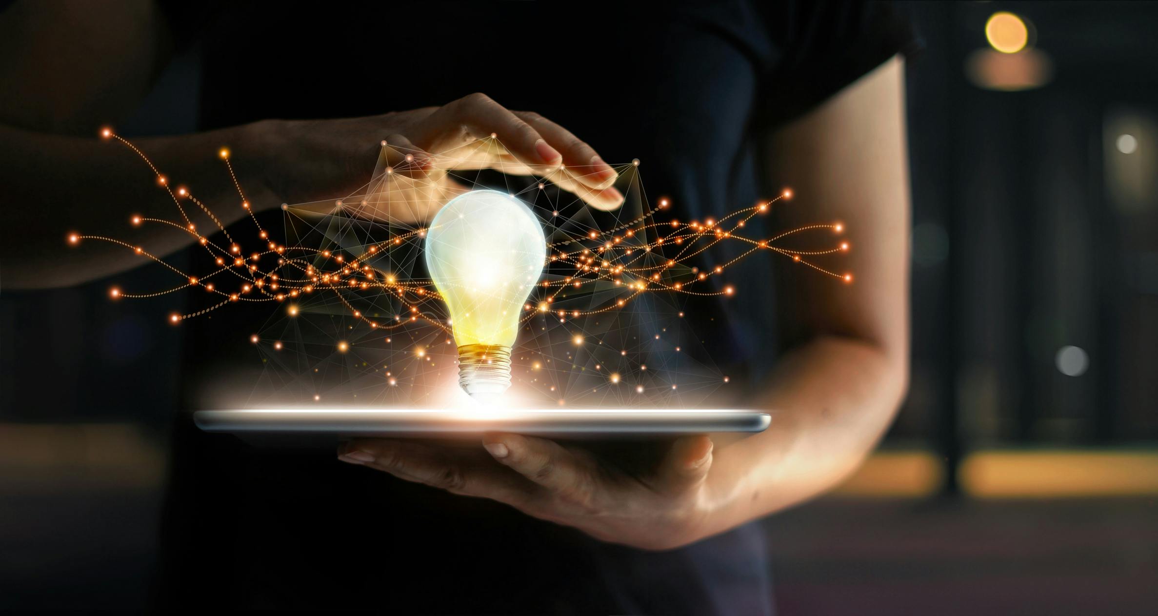Hands holding tablet with light bulb hovering over it, on virtual interface background, representing innovation.