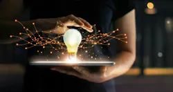 Hands holding tablet with light bulb hovering over it, on virtual interface background, representing innovation.