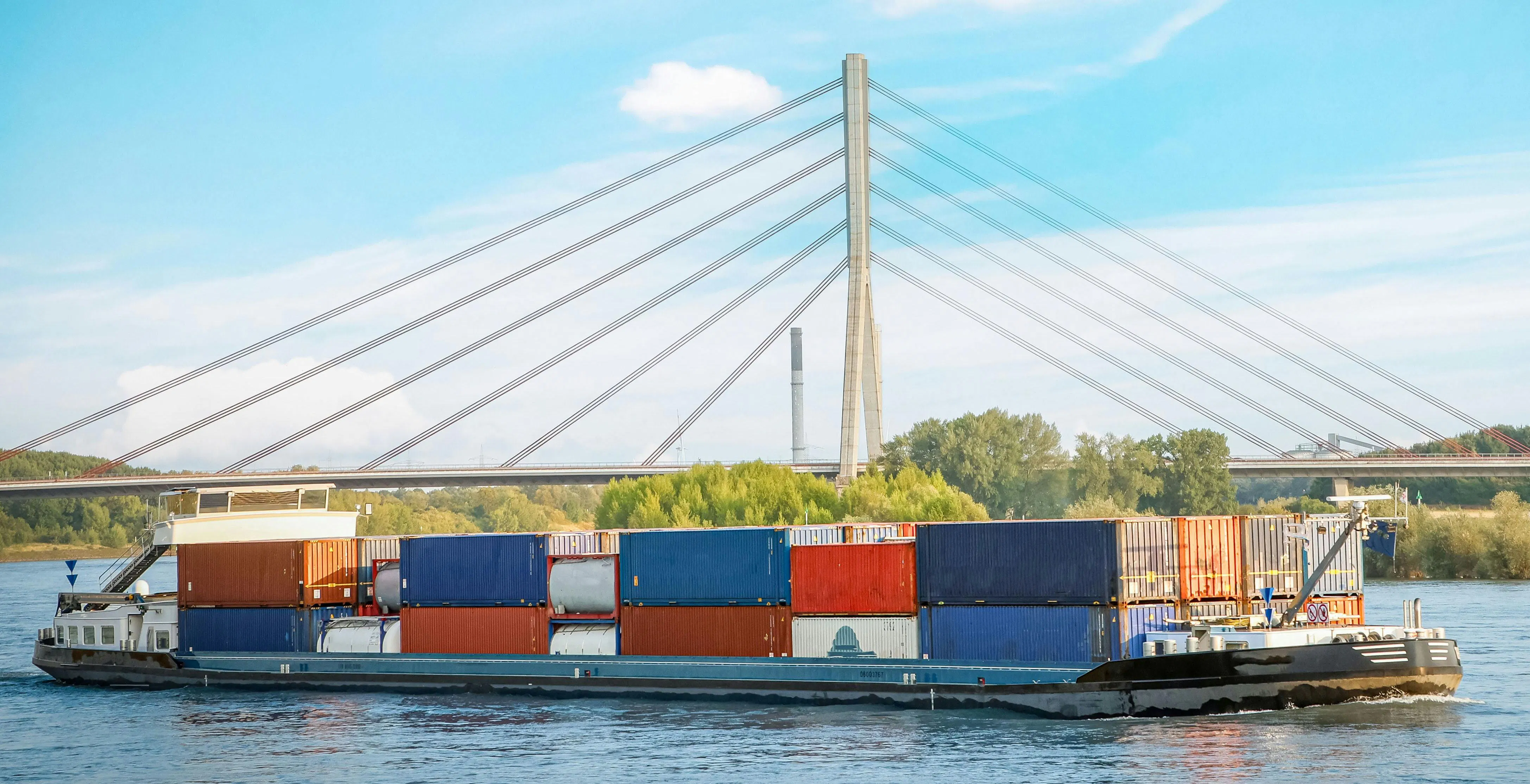 Cargo barge loaded with shipping containers sailing on the Rhine River, passing under a modern cable-stayed bridge.